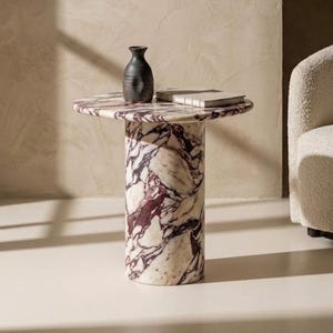 Könnte beinhalten: Ein runder Beistelltisch aus Marmor mit zylindrischem Sockel. Der Tisch hat ein weißes und burgunderfarbenes Muster. Eine dunkle Vase und ein Buch liegen auf der Tischplatte. Der Tisch ist etwa 61 cm hoch.
