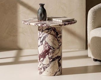May include: A round marble side table with a cylindrical base. The table has a white and burgundy pattern. A dark vase and a book rest on the tabletop. The table is approximately 61 cm tall.