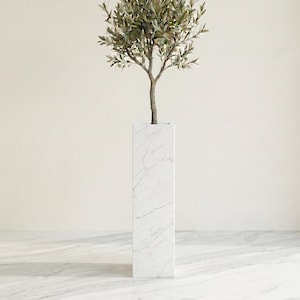 Könnte beinhalten: Ein hoher, rechteckiger weißer Marmor-Pflanzkübel mit einem künstlichen Olivenbaum. Der Pflanzkübel hat ein geädertes Muster und ein minimalistisches Design. Der Baum hat grüne Blätter und kleine olivenartige Früchte.