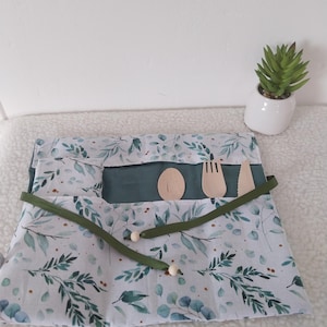 Può includere: Un portaposate in tessuto con un motivo di foglie di eucalipto verdi e verde acqua su sfondo bianco. Il portaposate contiene tre utensili in legno: un cucchiaio, una forchetta e un coltello, fissati da un nastro verde scuro con due perline.