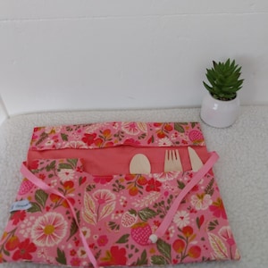 Op de afbeelding: Een roze stoffen bestekrol met een bloemen- en aardbeienpatroon, voorzien van drie binnenvakken met een houten lepel, vork en mes. De rol wordt vastgemaakt met twee roze linten die aan de uiteinden zijn voorzien van kleine houten kralen.