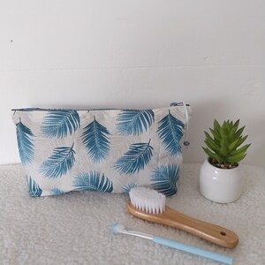 Può includere: Un beauty case bianco con un motivo a foglie di palma blu, una spazzola di legno con setole bianche e un pennello per il trucco azzurro. È visibile anche una piccola pianta grassa in vaso.