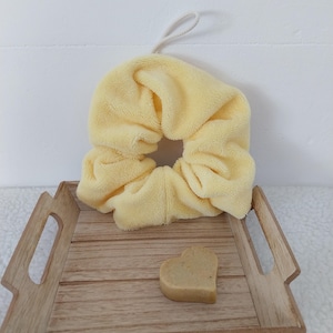 Puede incluir: Un coletero de tela amarillo pálido con un pequeño lazo blanco, colocado sobre una bandeja de madera marrón claro junto a un pequeño objeto sólido en forma de corazón de color tostado con superficie texturizada y asas recortadas.
