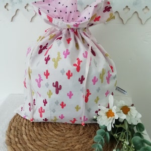 Peut inclure: Un sac blanc à cordon de serrage avec une doublure rose à motifs de cœurs. L'extérieur présente un motif répété de cactus colorés en rouge, rose, jaune et gris. Le sac est noué avec un ruban blanc et repose sur un tapis tissé.