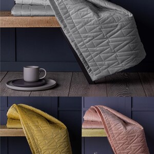 May include: A grey quilted throw blanket with a geometric design is displayed on a wooden shelf. A matching mug and plates are also on the shelf. Additional throws in yellow and pink are shown. The blankets are made of a soft material.