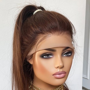May include: A brown human hair wig with a long, straight style. The wig is pulled back into a high ponytail and secured with a hair tie. The wig is worn on a mannequin head with a nude-colored wig cap. The mannequin is wearing a gold chain necklace with a crescent moon pendant.
