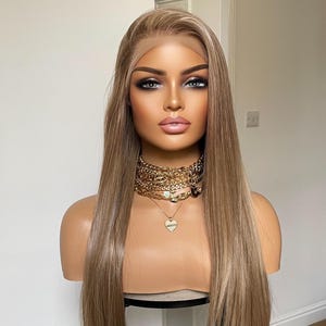 May include: A mannequin head displaying a long, straight, light brown wig with a lace front. The mannequin wears a gold chain necklace with a heart pendant. The mannequin has full makeup, including smoky eye shadow and pink lipstick.
