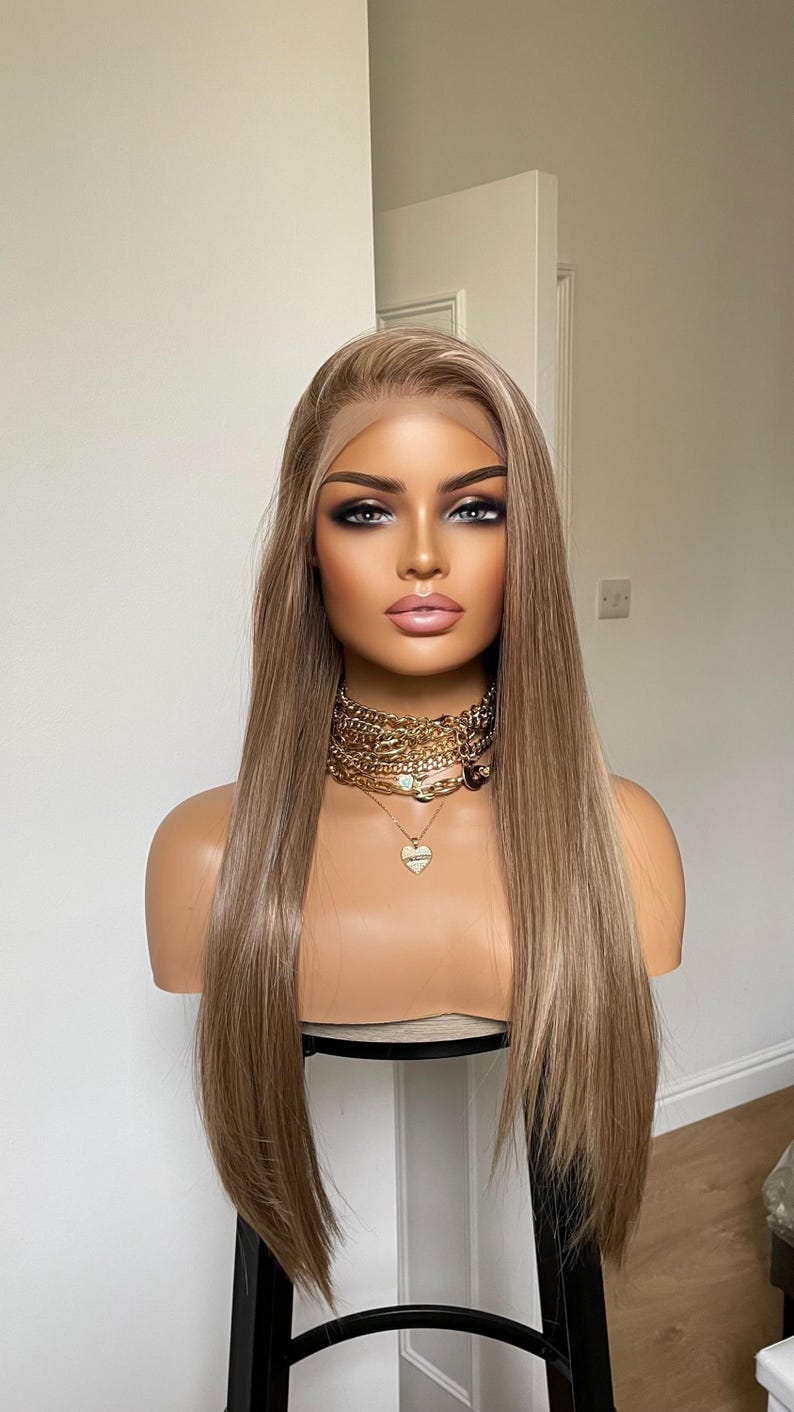 May include: A mannequin head displaying a long, straight, light brown wig with a lace front. The mannequin wears a gold chain necklace with a heart pendant. The mannequin has full makeup, including smoky eye shadow and pink lipstick.