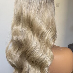 Puede incluir: Peluca rubia con ra&iacute;ces oscuras y cabello largo y ondulado. La peluca est&aacute; peinada con rizos voluminosos y se muestra en una cabeza de maniqu&iacute;. El color del cabello es una mezcla de rubio claro y marr&oacute;n m&aacute;s oscuro.