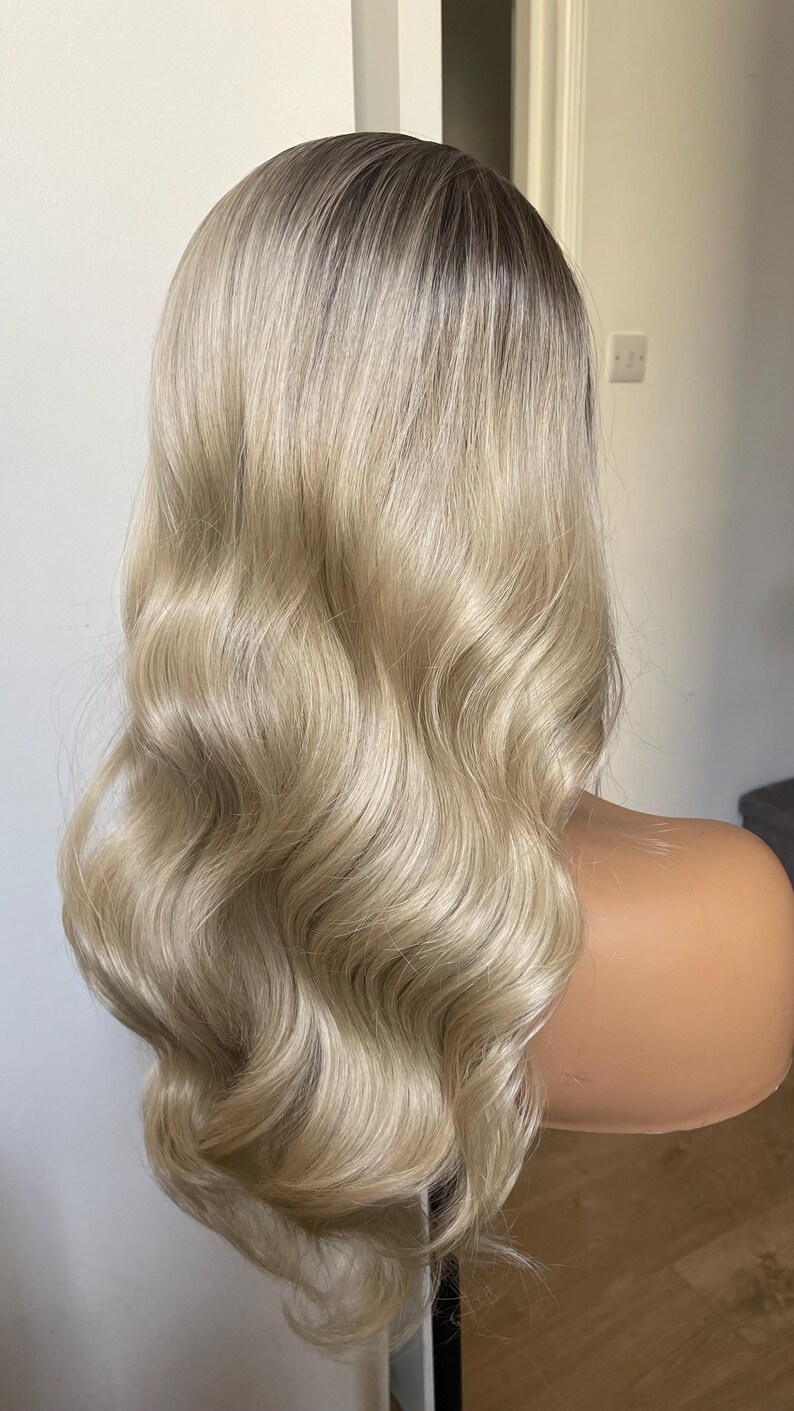 Puede incluir: Peluca rubia con ra&iacute;ces oscuras y cabello largo y ondulado. La peluca est&aacute; peinada con rizos voluminosos y se muestra en una cabeza de maniqu&iacute;. El color del cabello es una mezcla de rubio claro y marr&oacute;n m&aacute;s oscuro.
