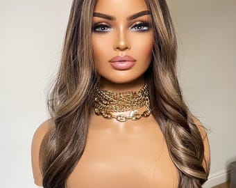 Op de afbeelding: Een lange, golvende bruine pruik met blonde highlights. De pruik is gestyled met losse golven en wordt gedragen op een mannequinhoofd. De mannequin draagt een gouden ketting.