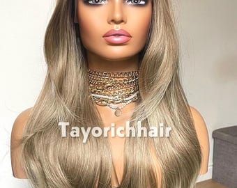 May include: A long, wavy, ash-blonde wig with dark roots on a mannequin head. The wig features a lace front and is styled with a center part. The mannequin wears a layered gold chain necklace and a pearl choker. The text Tayorichhair is visible.