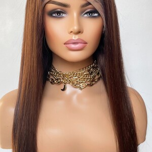 May include: A long, straight, brown human hair wig with a lace front. The wig is styled with a center part and falls to the shoulders. The wig is worn on a mannequin head with a gold chain necklace.