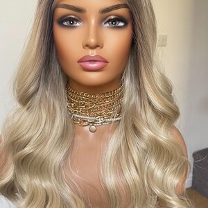 Puede incluir: Peluca rubia con capas onduladas y ra&iacute;ces oscuras. La peluca est&aacute; peinada sobre una cabeza de maniqu&iacute; con un rostro maquillado. Un collar de cadena dorada est&aacute; alrededor del cuello.