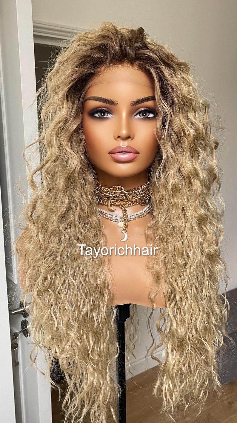 K&ouml;nnte beinhalten: Eine lange, blonde Per&uuml;cke mit lockeren Locken. Die Per&uuml;cke ist mit einem Mittelscheitel gestylt und f&auml;llt bis zu den Schultern. Die Per&uuml;cke sitzt auf einem Mannequin-Kopf mit einer goldenen Halskette.