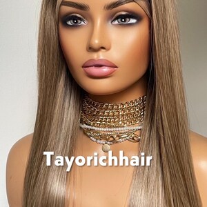 May include: A mannequin head with long, straight, light brown hair. The hair has subtle highlights. The mannequin wears a gold chain necklace and a choker. The text "Tayorichhair" is displayed at the bottom.