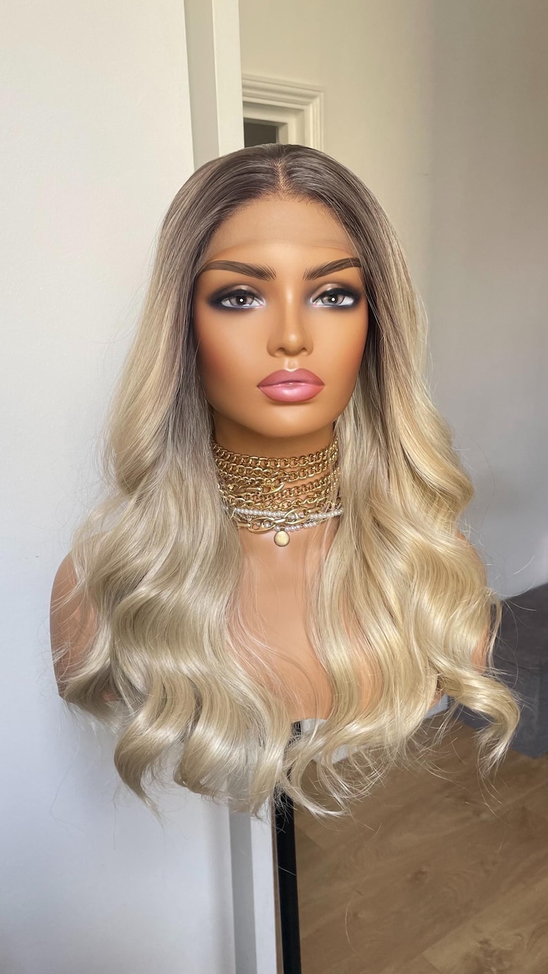 Puede incluir: Una peluca rubia con ra&iacute;ces oscuras peinada en una cabeza de maniqu&iacute;. La peluca presenta un cabello largo y ondulado de color rubio claro. El maniqu&iacute; lleva un collar de cadena dorada. El fondo es una pared blanca lisa.
