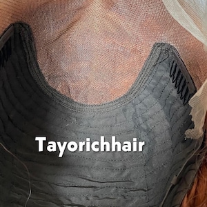 May include: Close-up of a wig cap with the text "Tayorichhair". The cap is dark gray with a curved design and small combs. The wig's lace front is visible, along with the reddish-brown hair.