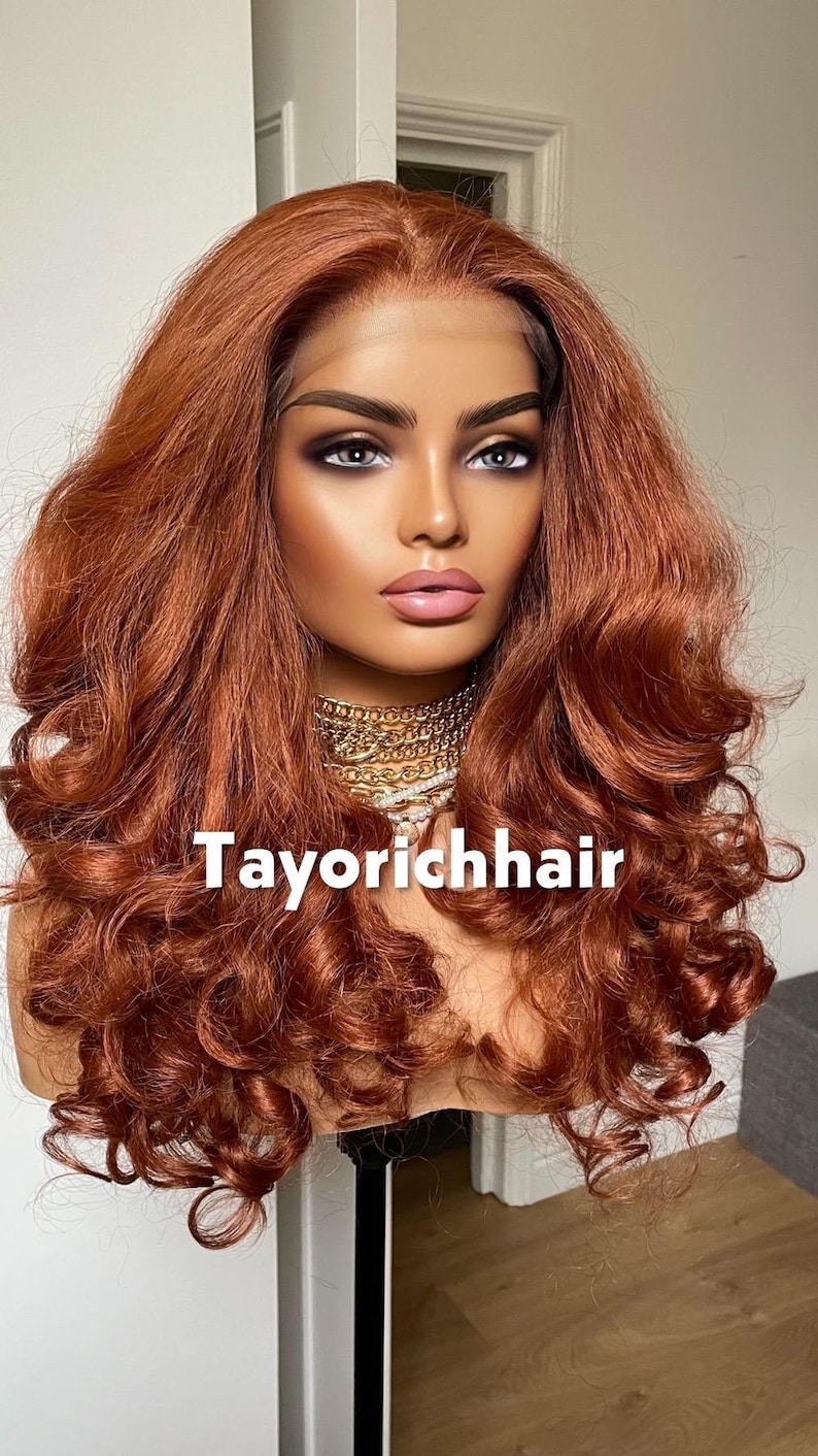 May include: A mannequin head displaying a long, wavy auburn wig. The wig features voluminous curls and a lace front. The text "Tayorichhair" is visible on the front. The model is wearing a gold necklace.