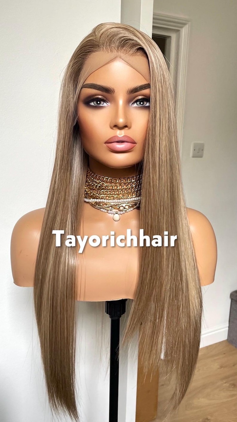 May include: A mannequin head displaying a long, straight blonde wig with subtle highlights. The wig is styled with a lace front and is on a stand. The text "Tayorichhair" is displayed on the mannequin.