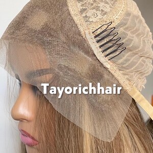 May include: A blonde wig with a lace front and the text "Tayorichhair" in white. The wig features a beige lace cap with black combs for secure attachment. The hair is a blend of blonde and light brown tones.