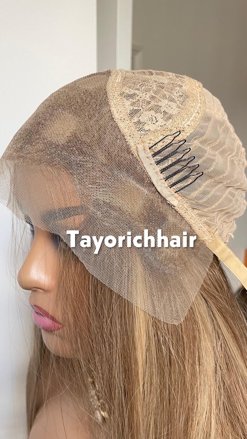 May include: A blonde wig with a lace front and the text "Tayorichhair" in white. The wig features a beige lace cap with black combs for secure attachment. The hair is a blend of blonde and light brown tones.