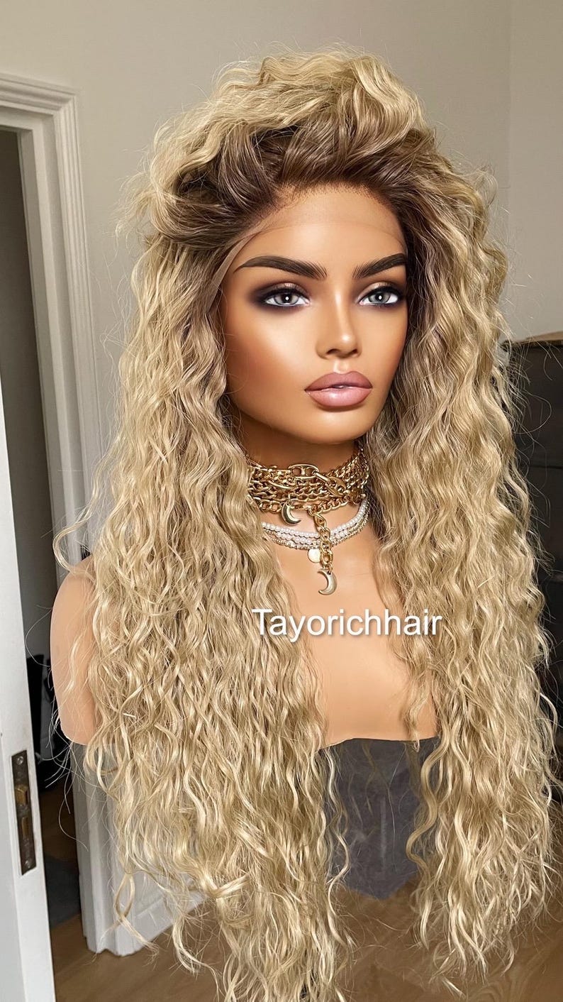 K&ouml;nnte beinhalten: Eine lange, blonde Per&uuml;cke mit lockeren, welligen Locken. Die Per&uuml;cke ist mit einem Mittelscheitel gestylt und hat einen nat&uuml;rlich aussehenden Haaransatz. Die Per&uuml;cke wird von einer Mannequin mit neutralem Gesichtsausdruck getragen.