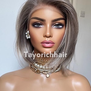 May include: A mannequin head wearing a short, straight, ash-blonde bob wig with a lace front. The mannequin features dark eyebrows, grey eyes, and pink lips. It is adorned with white flower earrings and layered gold and pearl necklaces. The text Tayorichhair is visible.