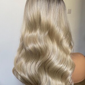 Puede incluir: Una peluca rubia larga y ondulada con ra&iacute;ces oscuras. La peluca est&aacute; peinada con ondas voluminosas y se muestra en una cabeza de maniqu&iacute;. El cabello tiene un degradado de color de aspecto natural y una textura brillante.