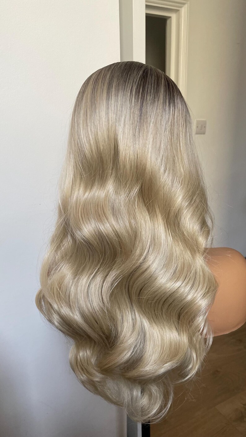 Puede incluir: Una peluca rubia larga y ondulada con ra&iacute;ces oscuras. La peluca est&aacute; peinada con ondas voluminosas y se muestra en una cabeza de maniqu&iacute;. El cabello tiene un degradado de color de aspecto natural y una textura brillante.