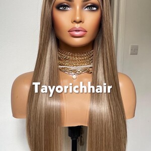 May include: A mannequin head displaying a long, straight wig with a blend of blonde and light brown highlights. The wig is styled with a middle part. The text "Tayorichhair" is displayed on the mannequin.