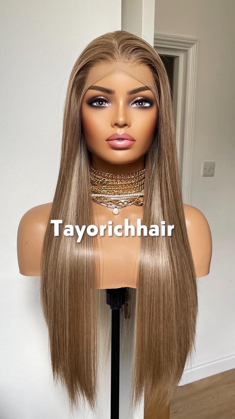 May include: A mannequin head displaying a long, straight wig with a blend of blonde and light brown highlights. The wig is styled with a middle part. The text "Tayorichhair" is displayed on the mannequin.