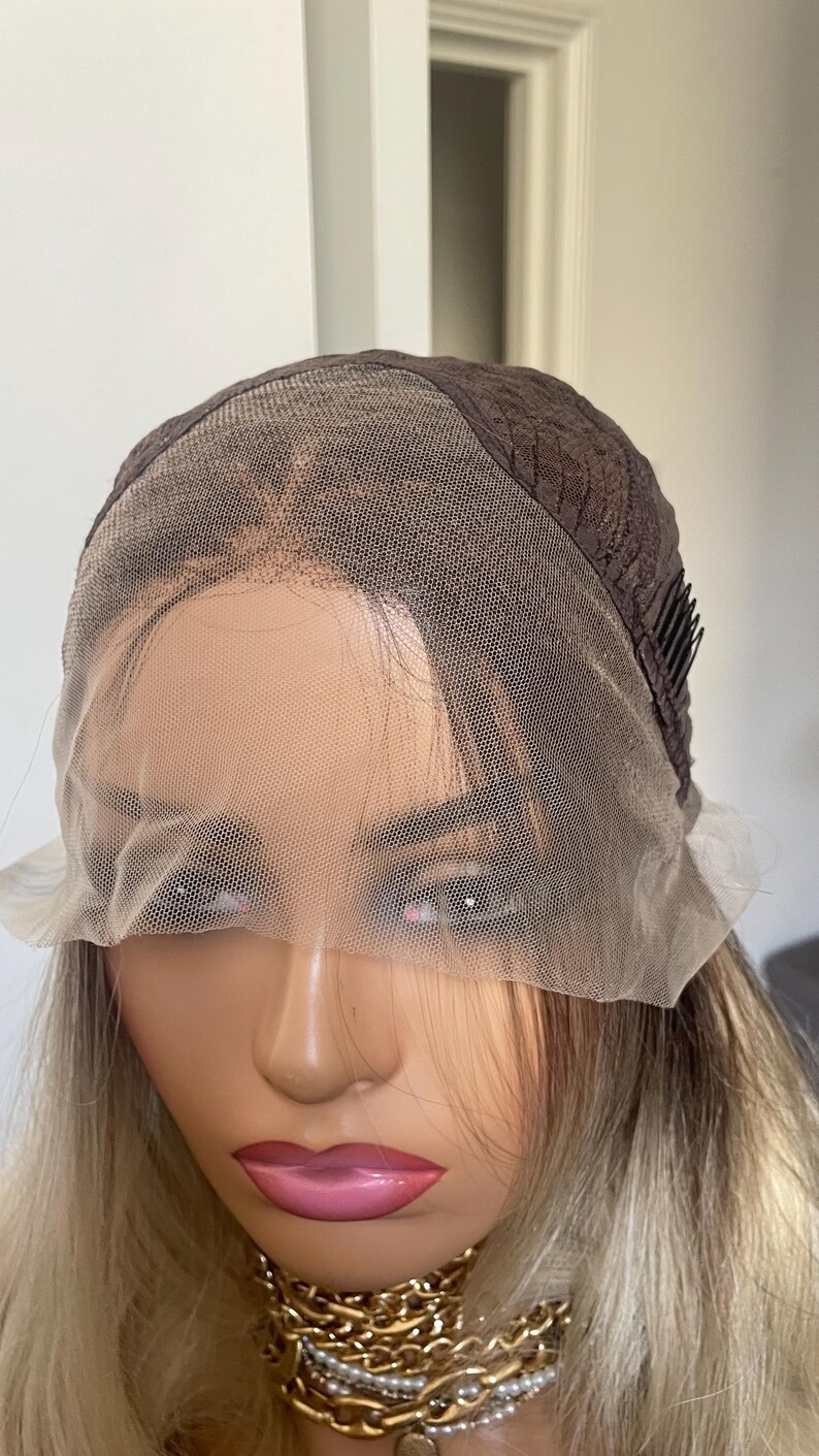 Puede incluir: Una peluca marr&oacute;n y rubia en una cabeza de maniqu&iacute;. La peluca presenta una parte delantera de encaje y una gorra marr&oacute;n oscuro. El cabello es una mezcla de tonos rubios y marrones. El maniqu&iacute; tiene labios rosados y un collar dorado.