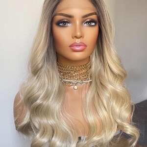 Puede incluir: Peluca rubia con ra&iacute;ces oscuras peinada sobre una cabeza de maniqu&iacute;. La peluca presenta un cabello largo y ondulado con un corte en capas. El maniqu&iacute; lleva un collar de cadena dorada. El fondo es una pared blanca lisa.