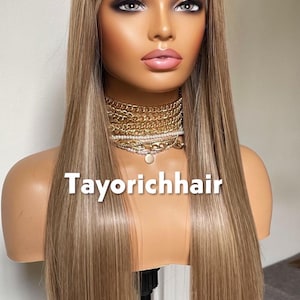 May include: A long, straight blonde wig with highlights is displayed on a mannequin head. The wig features a lace front and is styled with a center part. The mannequin is wearing a gold necklace. The text "Tayorichhair" is printed on the mannequin.