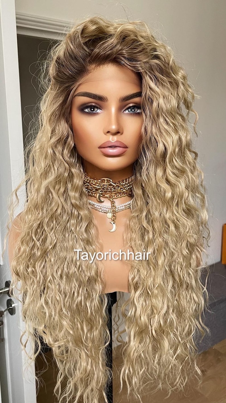 K&ouml;nnte beinhalten: Eine lange, blonde Per&uuml;cke mit lockeren, welligen Locken. Die Per&uuml;cke ist auf einem Mannequin-Kopf gestylt und hat einen nat&uuml;rlich aussehenden Haaransatz. Die Per&uuml;cke ist mit einem Mittelscheitel gestylt und f&auml;llt bis zu den Schultern. Die Per&uuml;cke ist mit "Tayorichhair" beschriftet.