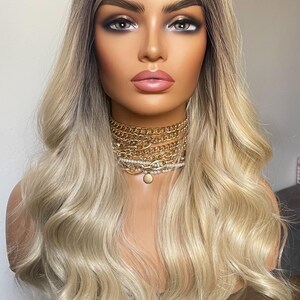 Puede incluir: Cabeza de maniqu&iacute; con peluca rubia con ra&iacute;ces oscuras y capas onduladas. El maniqu&iacute; lleva un collar de cadena dorada en capas. La cara est&aacute; completamente maquillada con sombra de ojos oscura y l&aacute;piz labial rosa.