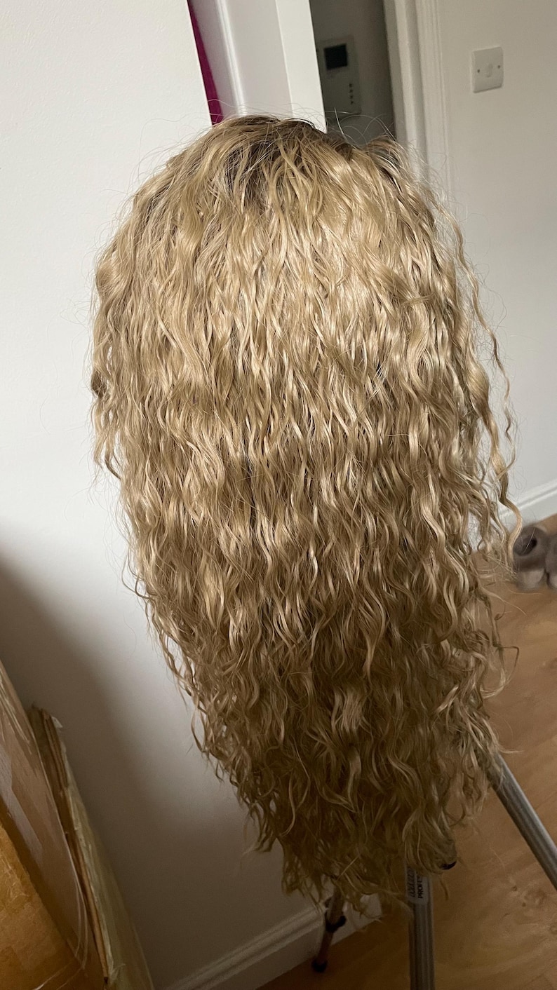 K&ouml;nnte beinhalten: Eine lange, blonde Per&uuml;cke mit lockeren, lockigen Wellen. Die Per&uuml;cke ist in einem nat&uuml;rlichen, flie&szlig;enden Stil gestylt.
