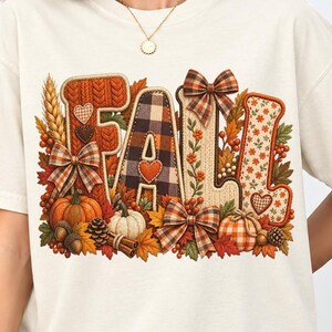 May include: An ivory short-sleeved t-shirt featuring a large graphic on the chest that spells the word FALL. The letters are decorated with autumn-themed patterns, including plaid, knit textures, pumpkins, bows, leaves, and small hearts.