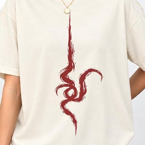 以下が含まれることがあります： アイボリー色の半袖Tシャツで、胸の中央に垂直な抽象的なダークレッドのグラフィックデザインが施されています。このデザインは、筆致のような流れる線で構成されており、ネックラインから胴体の中央に向かって曲線を描きながら伸びています。