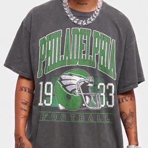 Può includere: Una t-shirt a maniche corte grigio antracite con una stampa grafica verde e bianca. Il testo recita PHILADELPHIA 1933 FOOTBALL, accompagnato dall'illustrazione di un casco da football. Il design presenta un aspetto vissuto e vintage.