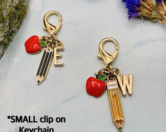 May include: Two gold-colored clip-on keychains. One features a red apple, a black and gold pencil, and the letter "E". The other has a red apple, a yellow and gold pencil, and the letter "W". The text "SMALL clip on Keychain 2 - 2 1/4" is at the bottom.