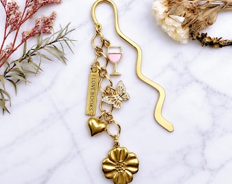 May include: A gold-toned metal bookmark with a wavy stem and a dangling charm cluster. The charms include a pink and gold wine glass, a rectangular tag reading I LOVE BOOKS, a crystal-accented butterfly, a gold heart, and a five-petal flower.