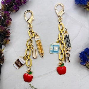 May include: Two gold-toned metal keychains featuring lobster clasps and chain links. The left keychain includes a coffee cup, a letter U, a yellow pencil, and a red apple. The right keychain features a blue book, a black pencil, a letter R, and a red apple.
