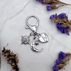 May include: Silver-toned keychain with a lobster clasp, featuring charms of a sunburst with a blue center, a crescent moon with stars, and a heart engraved with the letter "A". The keychain is displayed on a marble surface with dried purple flowers.