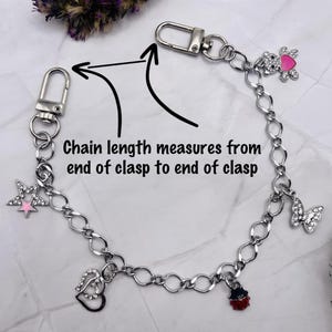 May include: A silver-toned metal chain featuring two lobster clasps at the ends and multiple charms including a star, double hearts, a ladybug, a butterfly, and a teddy bear with a pink heart. Text reads: Chain length measures from end of clasp to end of clasp.