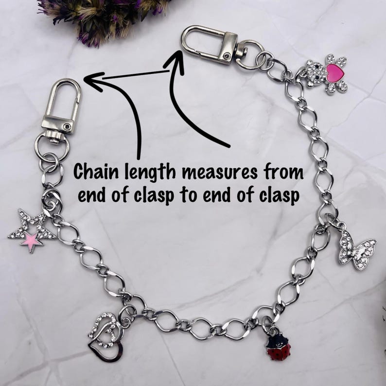 May include: A silver-toned metal chain featuring two lobster clasps at the ends and multiple charms including a star, double hearts, a ladybug, a butterfly, and a teddy bear with a pink heart. Text reads: Chain length measures from end of clasp to end of clasp.