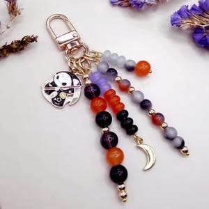 May include: A keychain featuring a gold-tone lobster clasp attached to a circular metal charm of a stylized character holding a scythe. Three dangling strands of beads in shades of orange, purple, black, and grey, accented with a gold crescent moon.