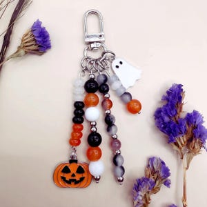 Puede incluir: Un llavero con un cierre de mosquet&oacute;n plateado y tres hilos colgantes de cuentas en naranja, negro, blanco y gris. Los hilos est&aacute;n adornados con un dije de calabaza de metal y un peque&ntilde;o dije de fantasma blanco.