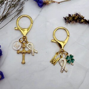 May include: Two gold-colored keychains with lobster claw clasps. One keychain has a cross, a white circle with the letter "M", and a rhinestone ribbon charm. The other has the letter "M", a rhinestone ribbon, and a green four-leaf clover charm.